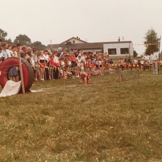 Eine Menschenmenge sieht zu, wie Kinder unter einem Reifen auf einem Grasfeld krabbeln. Im Hintergrund sind Gebäude und Bäume zu sehen.