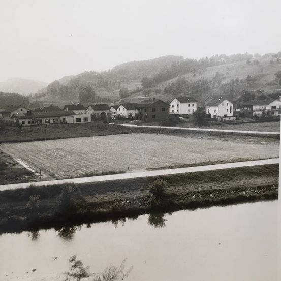 Ein Schwarz-Weiß-Foto einer ländlichen Landschaft mit Häusern am Hang und einem Fluss im Vordergrund.