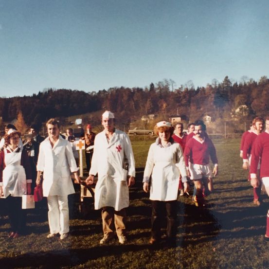 Eine Gruppe von Menschen in weißen Kitteln und Mützen, möglicherweise medizinisches Personal, geht auf einem Feld mit anderen in roten Trikots.
