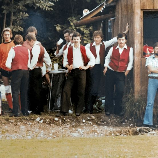 Eine Gruppe von Männern in roten Westen und weißen Hemden versammelt sich vor einer Holzstruktur, einige halten Tassen. Ein Mann steht mit seinem Arm auf der Wand.