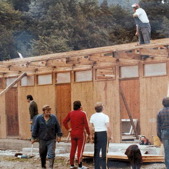 Mehrere Personen arbeiten an einem Holzhaus im Bau. Eine Person ist auf dem Dach, andere stehen oder gehen herum. Es gibt Holzbalken, Werkzeuge und einen gelben Eimer in der Nähe.