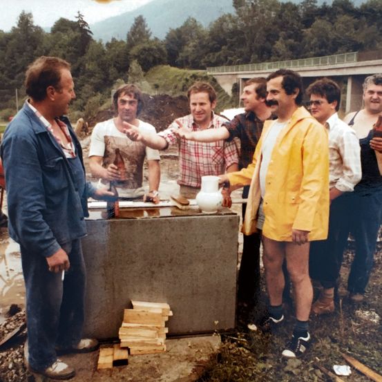 Eine Gruppe von Männern versammelt sich um einen provisorischen Tisch im Freien, einer hält eine Flasche und ein anderer eine Kanne. Die Umgebung ist ländlich mit Bäumen und einer Brücke im Hintergrund.