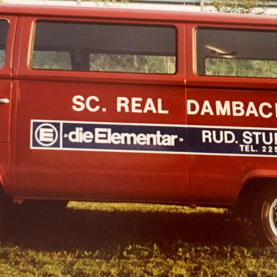 Ein roter Van mit weißen und blauen Aufklebern, die SC. REAL DAMBACH und dieElementar anzeigen, mit Rud. Stud. Tel. 225.