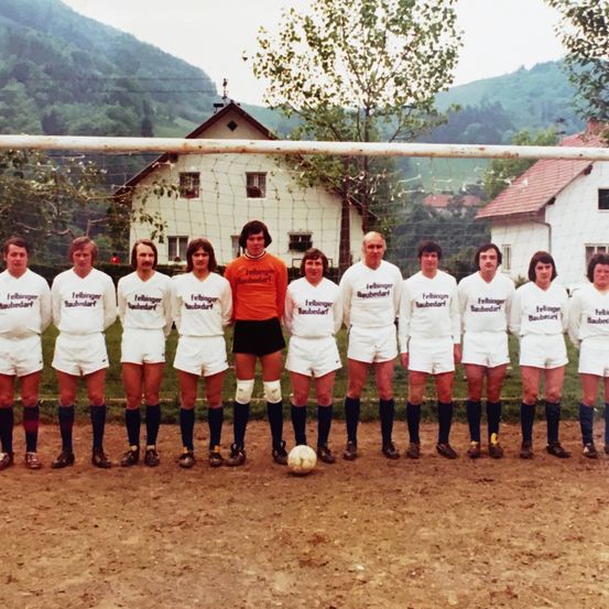 Ein Vintage-Foto einer Fußballmannschaft, die vor einem Tor steht, mit einem Fußball an ihren Füßen. Die Teammitglieder tragen weiße Uniformen mit Namen und Nummern.