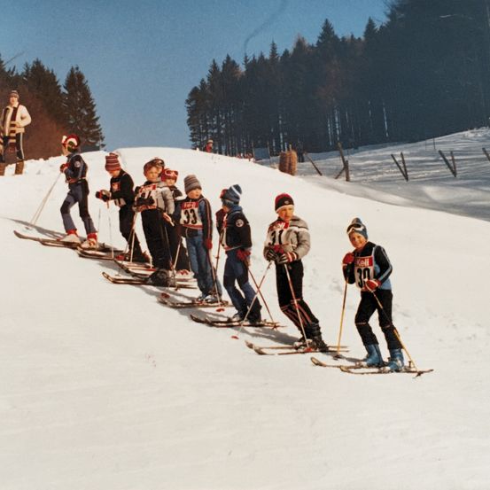 Eine Gruppe von Skifahrern, alle mit Nummern auf ihren Jacken, stehen in einer Reihe im Schnee. Sie halten Skistöcke und tragen Skibrillen. Ein Mann steht hinter ihnen im Schnee. Im Hintergrund befinden sich Kiefernbäume.