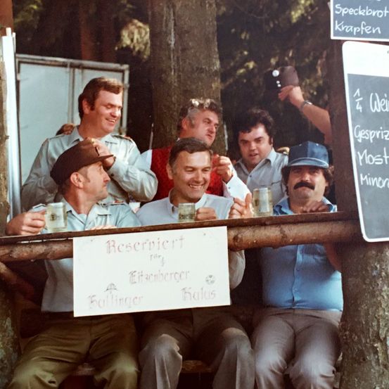 Eine Gruppe von Männern sitzt auf einer Holzbank, lächelt und hält Gläser mit Bier in der Hand. Sie scheinen auf einem Festival zu sein. Ein Schild steht 'Reserviert für Ettenberger Kolling'. Hinter ihnen zeigt ein Menübrett verschiedene Essensoptionen.