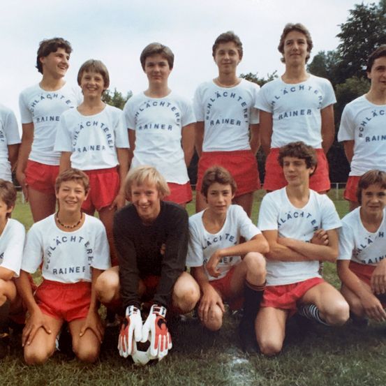 Ein Gruppenfoto einer Fußballmannschaft in den 1980er Jahren. Sie tragen passende weiße Trikots mit dem Aufdruck 'Rainer'. Eine Person kniet und hält einen Fußball.