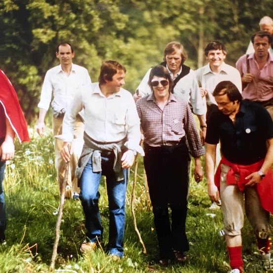 Eine Gruppe von Männern, in Freizeitkleidung gekleidet, geht durch ein Grasfeld. Ein Mann trägt eine Sonnenbrille und hält einen Wanderstock.