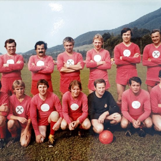 Eine Gruppe von Fußballspielern in roten Uniformen posiert für ein Foto auf einem Rasenfeld mit Bergen im Hintergrund.