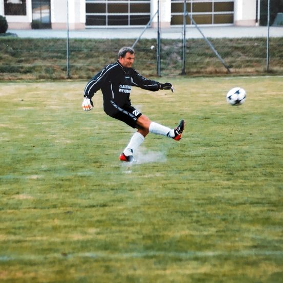 Ein Fußballspieler in einem schwarzen Trikot tritt einen Ball auf einem Rasenfeld. Der Ball ist in der Luft.
