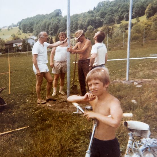 Fünf Männer arbeiten an einem Pfosten auf einem Feld, während ein Junge zusieht. Ein Mann zeigt auf den Pfosten, während ein anderer eine Schaufel hält.