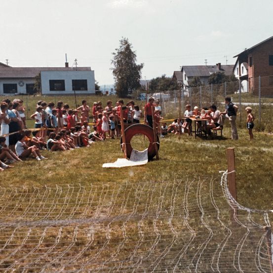 Ein altes Foto zeigt eine Gruppe von Menschen, die sich für eine Gemeinschaftsveranstaltung auf einer Wiese versammelt haben. Einige sitzen, andere stehen, mit einem roten Hindernis in der Mitte. Im Hintergrund sind Häuser und ein Zaun zu sehen.