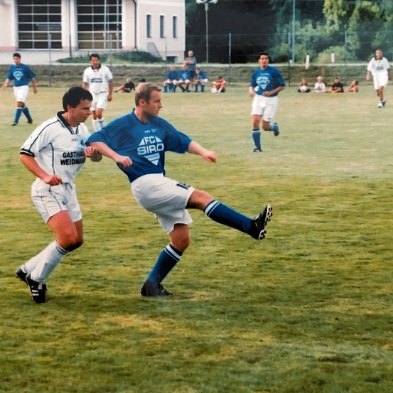 Zwei Fußballspieler spielen auf einem Feld. Einer ist in blau und weiß gekleidet, der andere in weiß und blau.