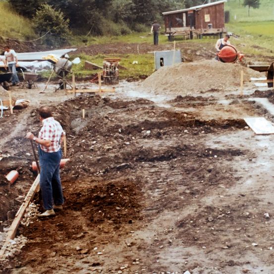 Eine Gruppe von Arbeitern befindet sich an einer Baustelle. Ein Mann gräbt mit einer Schaufel eine Grube.