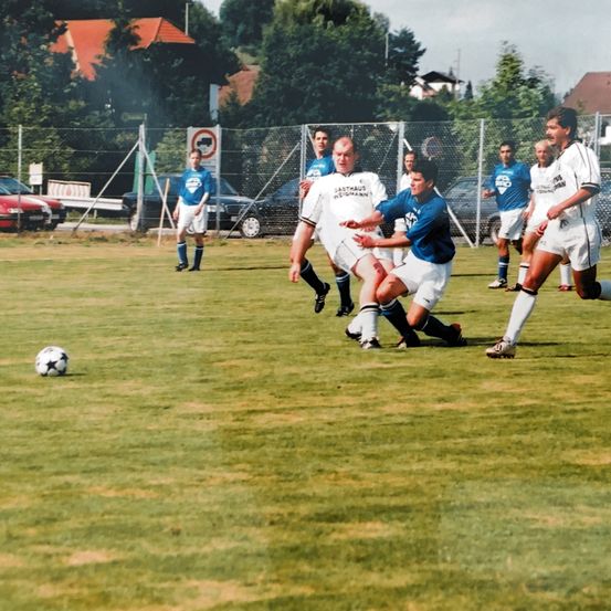 Fußballspieler in weißen und blauen Trikots spielen auf einem Feld. Ein Spieler in Blau tackelt einen Spieler in Weiß in der Nähe des Balls.