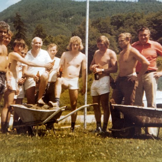 Eine Gruppe von Männern in Shorts und einigen in Hemden, die Flaschen halten, stehen um eine mit Erde gefüllte Schubkarre herum, mit einem Pfahl und einem Berg im Hintergrund.