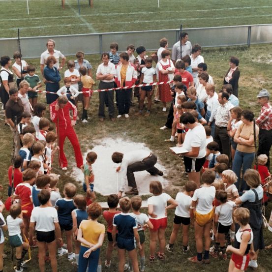 Eine Gruppe von Menschen versammelt sich auf einem Feld und beobachtet einen Mann, der eine Sandgrube misst. Der Mann wird von Kindern umringt, während andere hinter einer Barriere stehen.