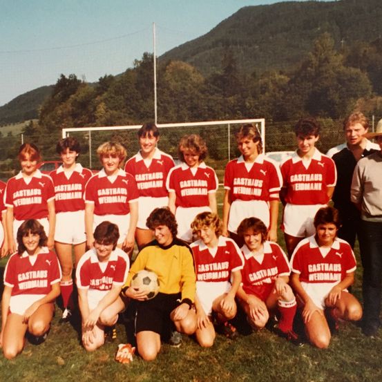 Eine Fußballmannschaft in roten und weißen Trikots, möglicherweise von Gasthaus, posiert für ein Foto auf einem Rasen mit Bergen im Hintergrund.