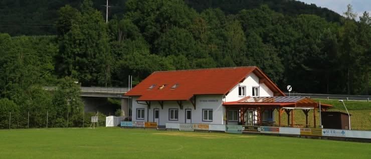 Ein kleines weißes Haus mit rotem Dach steht inmitten eines grünen Feldes. In der Nähe befindet sich eine Brücke, und im Hintergrund erhebt sich ein bewaldeter Berg.