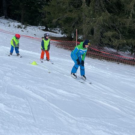 Drei Personen sind auf einem verschneiten Hang beim Skifahren, wobei eine Person führt und zwei hinterher sind. Sie tragen alle Winterkleidung und Helme. Der Boden ist mit Schnee bedeckt, und es gibt Bäume und ein Netz im Hintergrund.