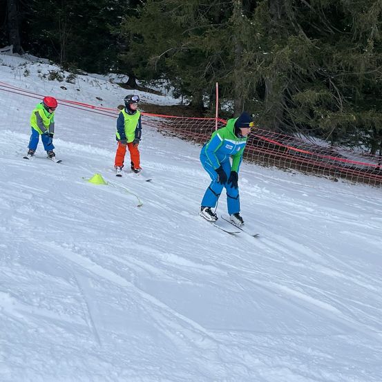 Drei Personen sind auf einem verschneiten Hang beim Skifahren, wobei eine Person führt und zwei hinterher sind. Sie tragen alle Winterkleidung und Helme. Der Boden ist mit Schnee bedeckt, und es gibt Bäume und ein Netz im Hintergrund.