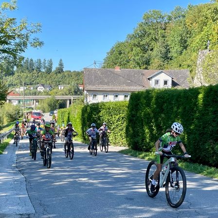 Eine Gruppe von Radfahrern fährt auf einer asphaltierten Straße, wobei mehrere Radfahrer Helme tragen. Im Hintergrund befindet sich ein Haus mit einem grauen Dach.
