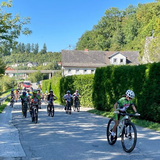 Eine Gruppe von Radfahrern fährt auf einer asphaltierten Straße, wobei mehrere Radfahrer Helme tragen. Im Hintergrund befindet sich ein Haus mit einem grauen Dach.