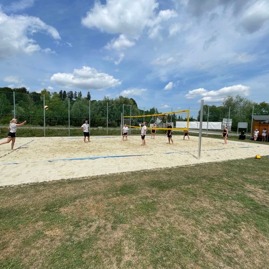 Eine Gruppe von Menschen spielt Volleyball auf einem sandigen Spielfeld mit einem Netz. Einige spielen aktiv, während andere zuschauen.