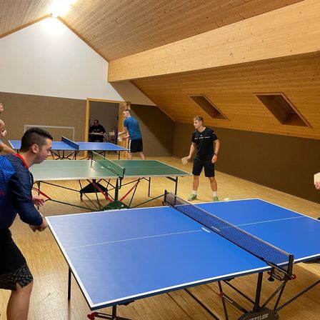 In einem hölzernen Raum spielen mehrere Personen Tischtennis. Ein Spieler ist kurz davor, den Ball zu schlagen. Ein anderer Spieler steht in der Nähe und beobachtet.