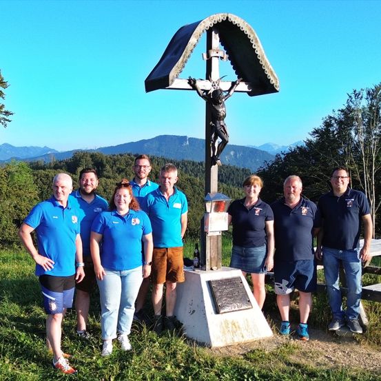 Eine Gruppe von Menschen in blauen Shirts und Shorts steht vor einem Kruzifix. Sie lächeln und scheinen auf einer Wanderung zu sein.