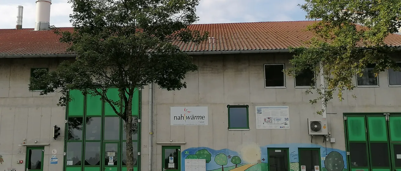 Ein großes Gebäude mit rotem Dach, grünen Türen und Wandgemälden. Vor dem Gebäude stehen Bäume und eine Bank.