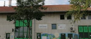 Ein großes Gebäude mit rotem Dach, grünen Türen und Wandgemälden. Vor dem Gebäude stehen Bäume und eine Bank.