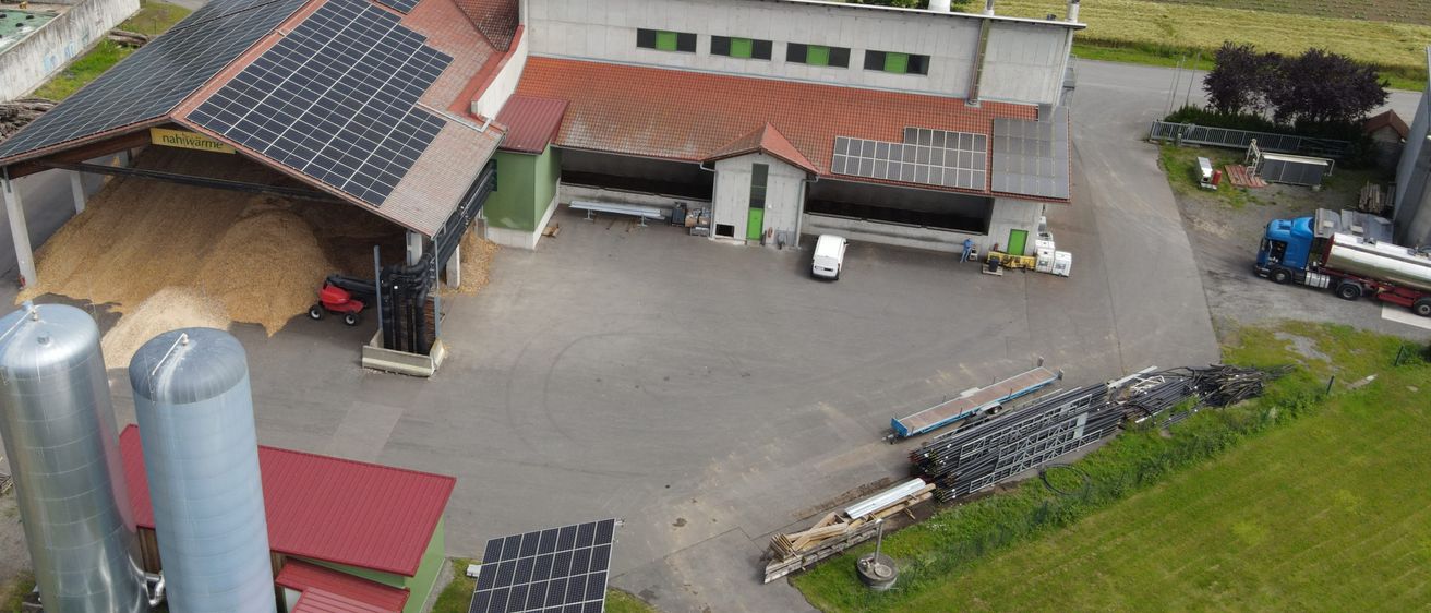 Eine Luftaufnahme einer Solaranlage mit Solarmodulen auf dem Dach und zwei großen Silos. In der Nähe befinden sich Gewächshäuser, Felder und ein Fluss.