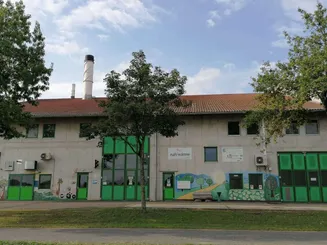 Ein Betongebäude mit rotem Dach und grünen Türen steht an einem klaren Tag. Davor steht ein Baum. Die Wand hat Wandbilder.