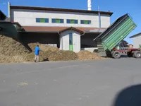 Ein Mann in einer blauen Jacke geht an einem Haufen Holzspäne vorbei, vor einem großen Gebäude mit Solarpanelen.
