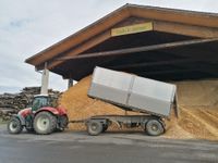 Ein roter Traktor entlädt Getreide in einen Anhänger mit einem Haufen Getreide dahinter unter einem Gebäude mit einem Schild, das 'nahwarme' steht.