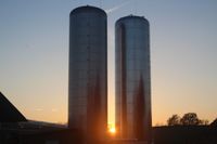 Zwei hohe zylindrische Metallsilos stehen nebeneinander, mit der untergehenden Sonne dahinter, die lange Schatten wirft.