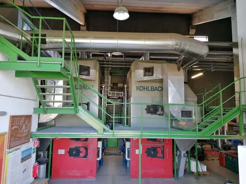 Eine Innenfabrik mit großen roten Kesseln, die mit 'KOHLBACH' und 'L3ACH' beschriftet sind. Die Fabrik hat Metalltreppen und Geländer, große Rohre und mehrere gestapelte Kisten.