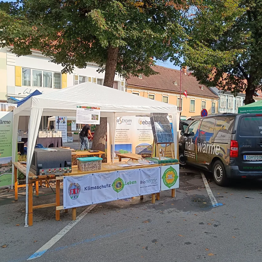 Ein weißes Zelt mit einem Banner zum Thema Klimaschutz steht auf einem Parkplatz. Eine Person steht daneben und ein Van mit Solarpanel ist geparkt.
