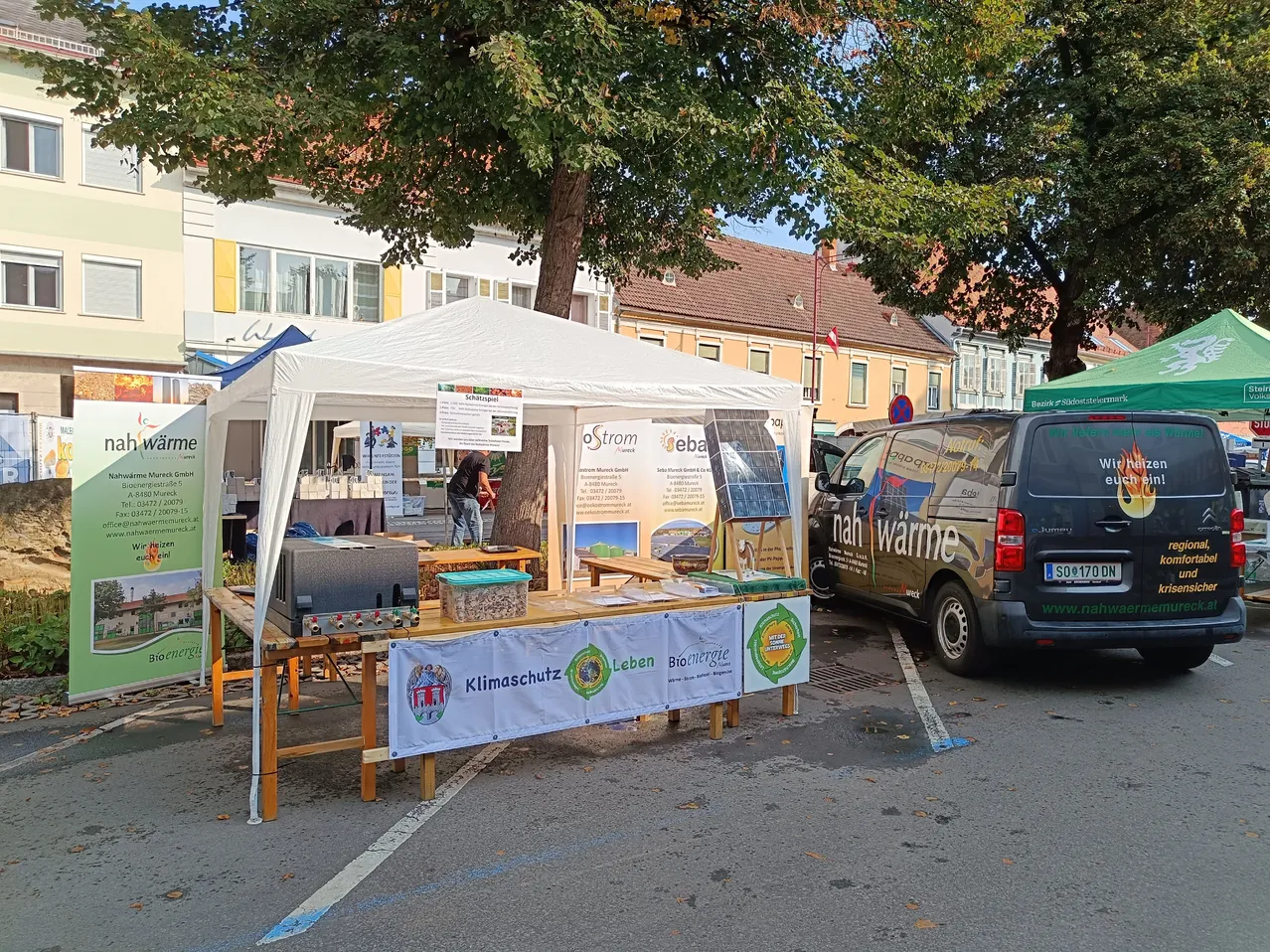 Ein weißes Zelt mit einem Banner zum Thema Klimaschutz steht auf einem Parkplatz. Eine Person steht daneben und ein Van mit Solarpanel ist geparkt.