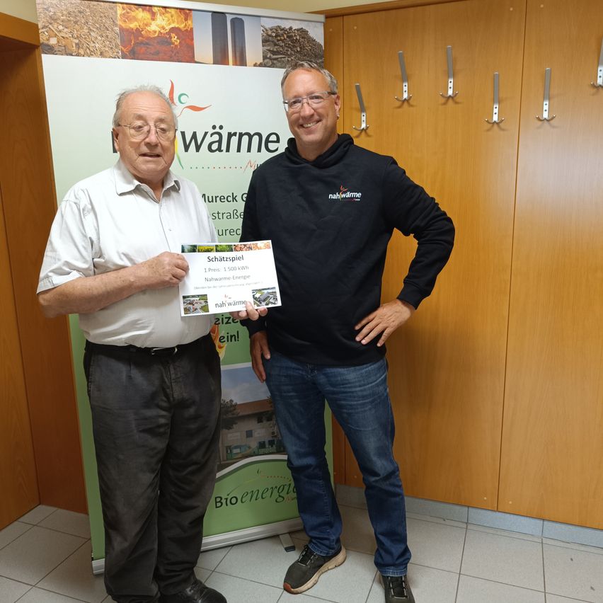 Zwei Männer stehen vor einem Schild mit einem Feuergrafik. Ein Mann hält ein Zertifikat. Sie tragen beide Freizeitkleidung und stehen vor einer Holzwand.