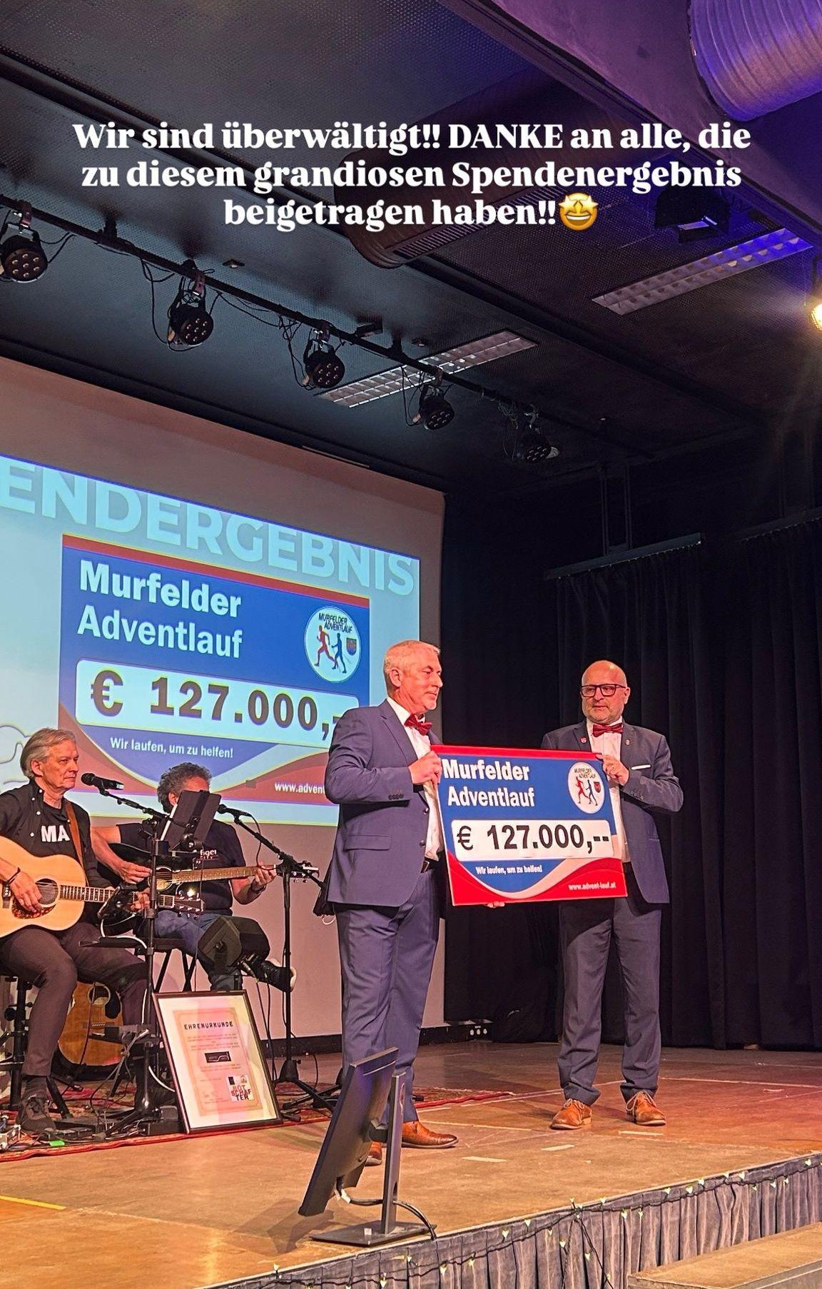 Zwei Männer halten ein großes Schild mit der Aufschrift Murfelder Adventlauf und einer großen Geldsumme. Ein Mann hält ein Mikrofon. Zwei Personen spielen Gitarren.
