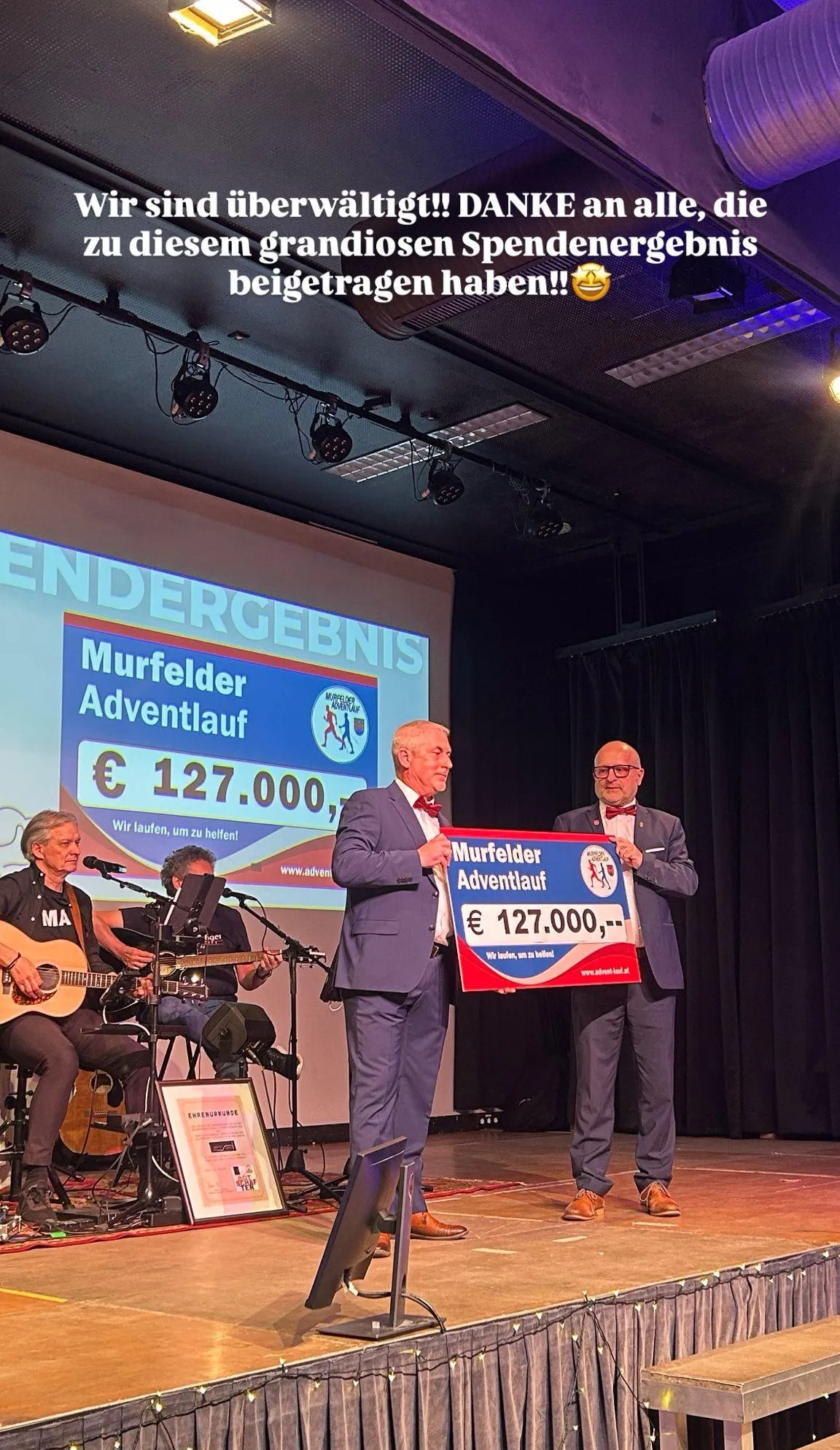 Zwei Männer halten ein großes Schild mit der Aufschrift Murfelder Adventlauf und einer großen Geldsumme. Ein Mann hält ein Mikrofon. Zwei Personen spielen Gitarren.
