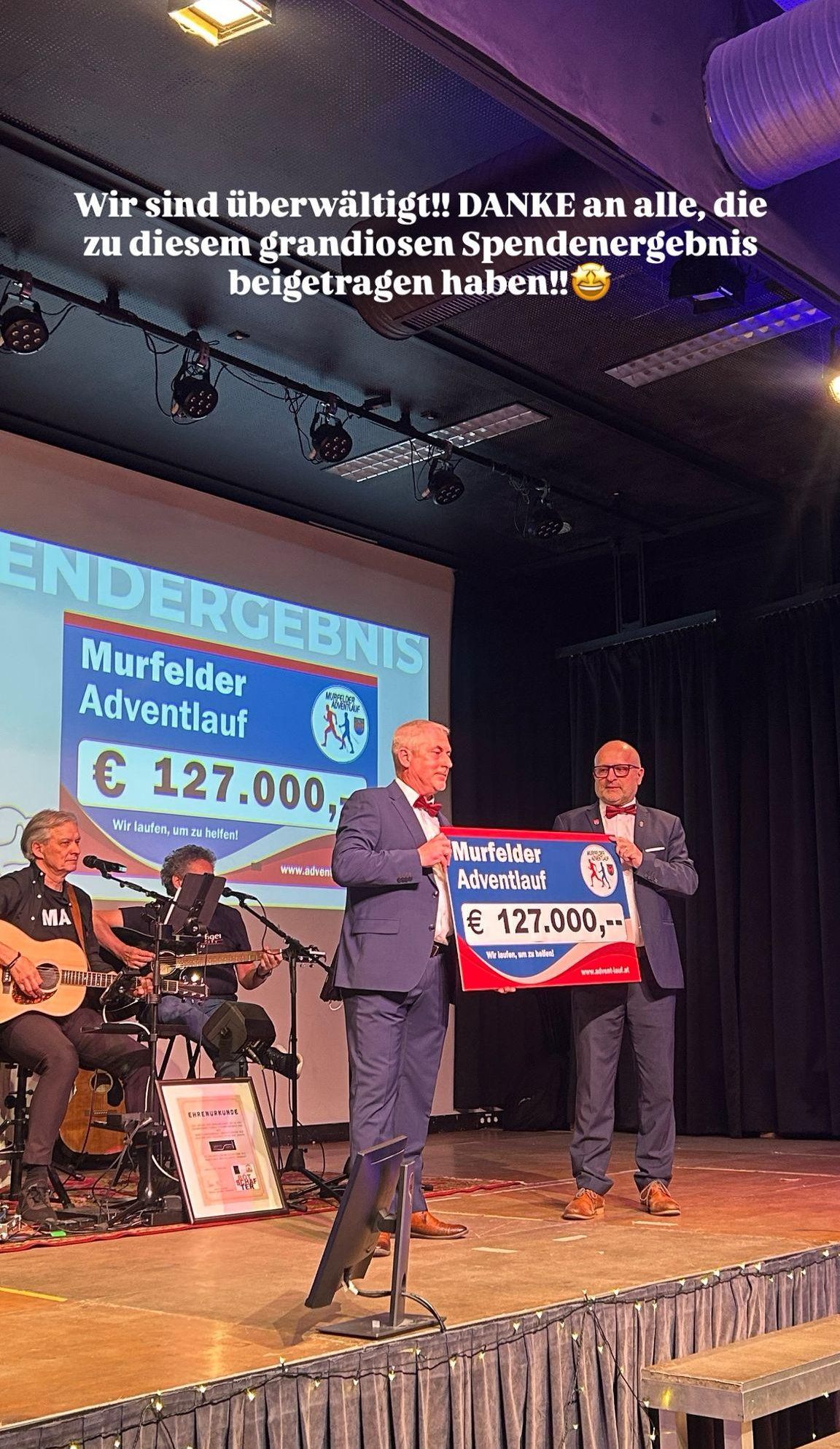 Zwei Männer halten ein großes Schild mit der Aufschrift Murfelder Adventlauf und einer großen Geldsumme. Ein Mann hält ein Mikrofon. Zwei Personen spielen Gitarren.