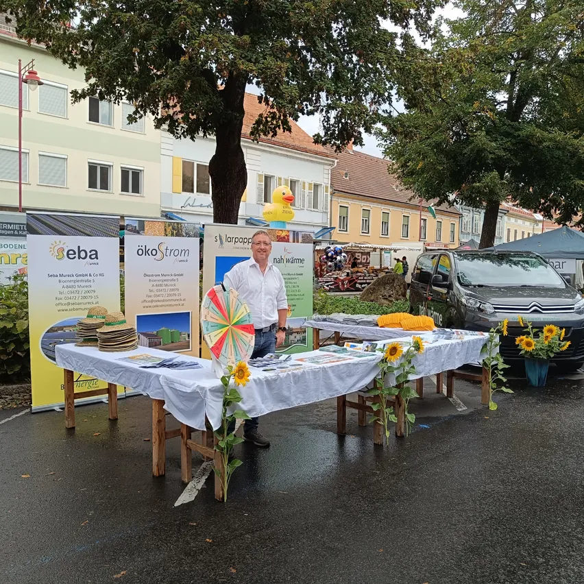 Ein Mann steht hinter einem Tisch mit Sonnenblumen und Werbematerialien auf einem Stadtplatz.