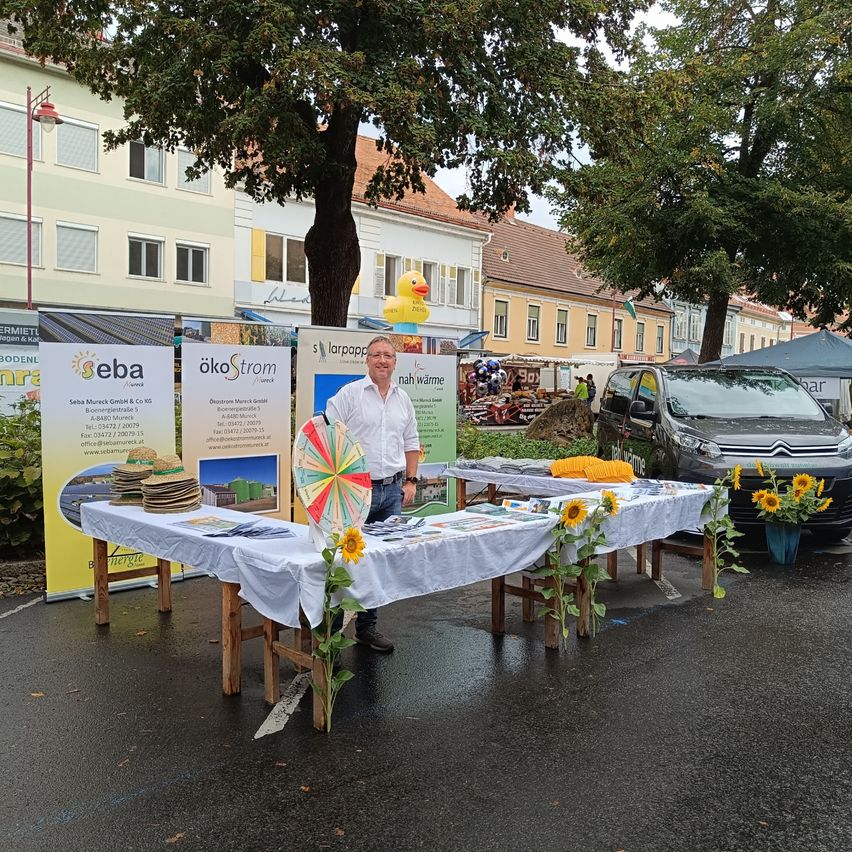 Ein Mann steht hinter einem Tisch mit Sonnenblumen und Werbematerialien auf einem Stadtplatz.