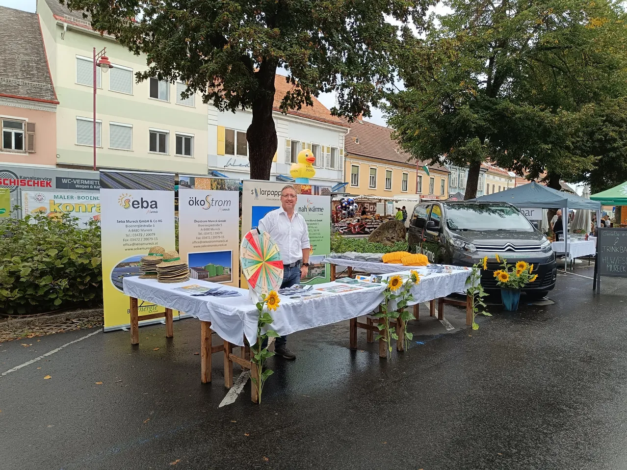 Ein Mann steht hinter einem Tisch mit Sonnenblumen und Werbematerialien auf einem Stadtplatz.