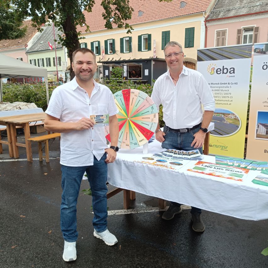 Zwei Männer stehen hinter einem Tisch mit Werbematerialien, einer hält eine Karte, in einer Außenumgebung mit Gebäuden und einem Zelt im Hintergrund.