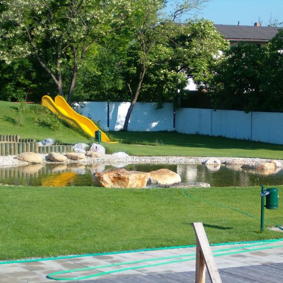 Ein Außenbereich mit einer gelben Rutsche, einem kleinen Teich mit Steinen, einer weißen Umzäunung und einem Mülleimer.