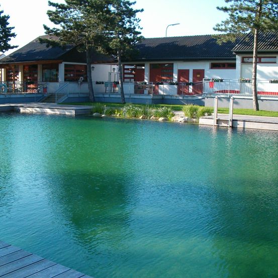 Ein modernes Gebäude mit schwarzem Dach und roten Türen steht neben einem ruhigen, grün-blauen Pool. Bäume und Pflanzen umgeben das Gebiet, und eine Straßenlaterne ist in der Ferne zu sehen.